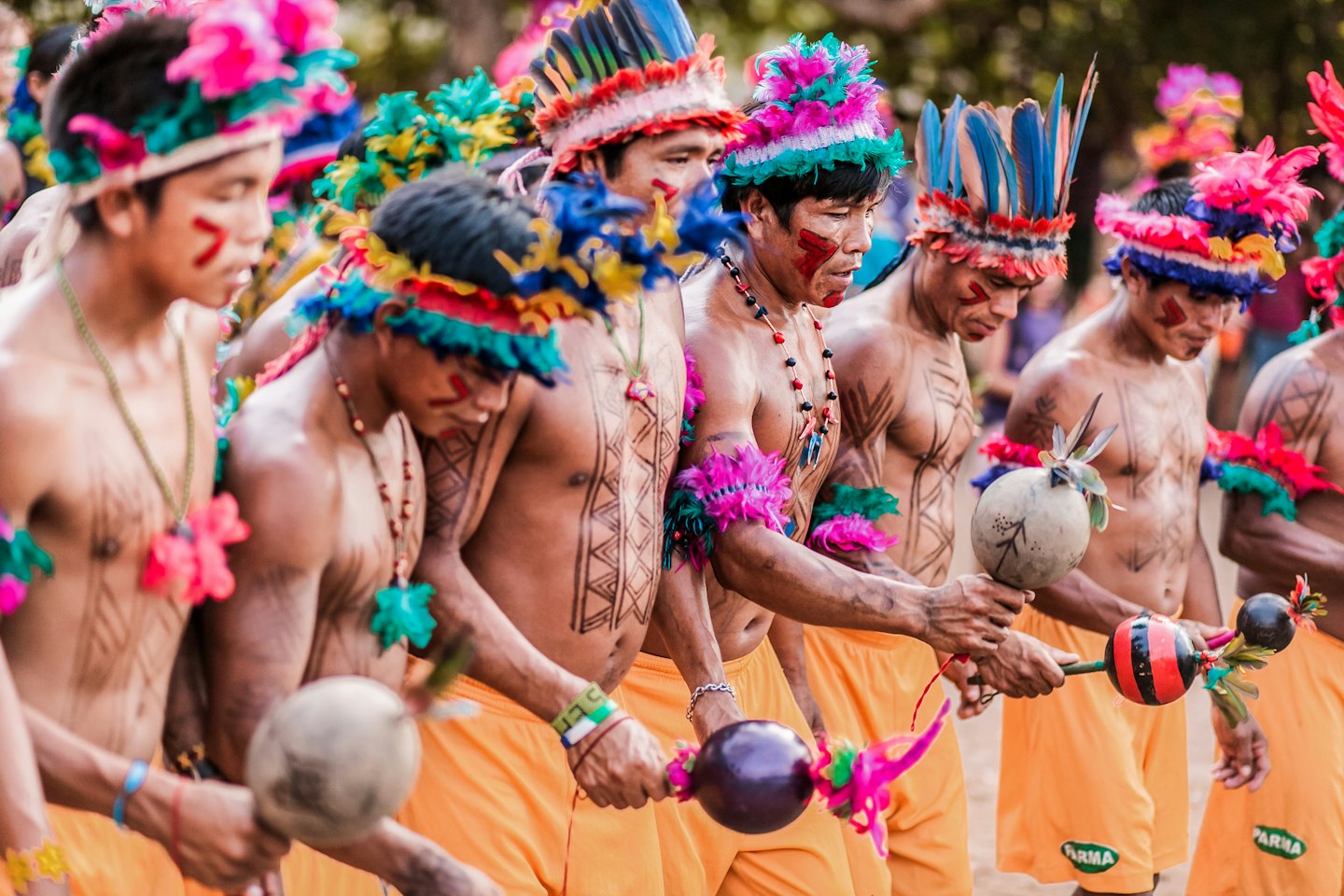 Indigenous men in colorful feathered headdresses perform a dance with maracas.